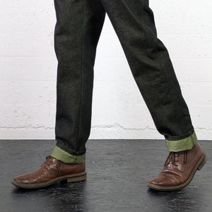 A person wearing dark charcoal gray textured trousers with a rolled hem showing olive green lining, styled with brown leather lace-up shoes against a white brick wall background.